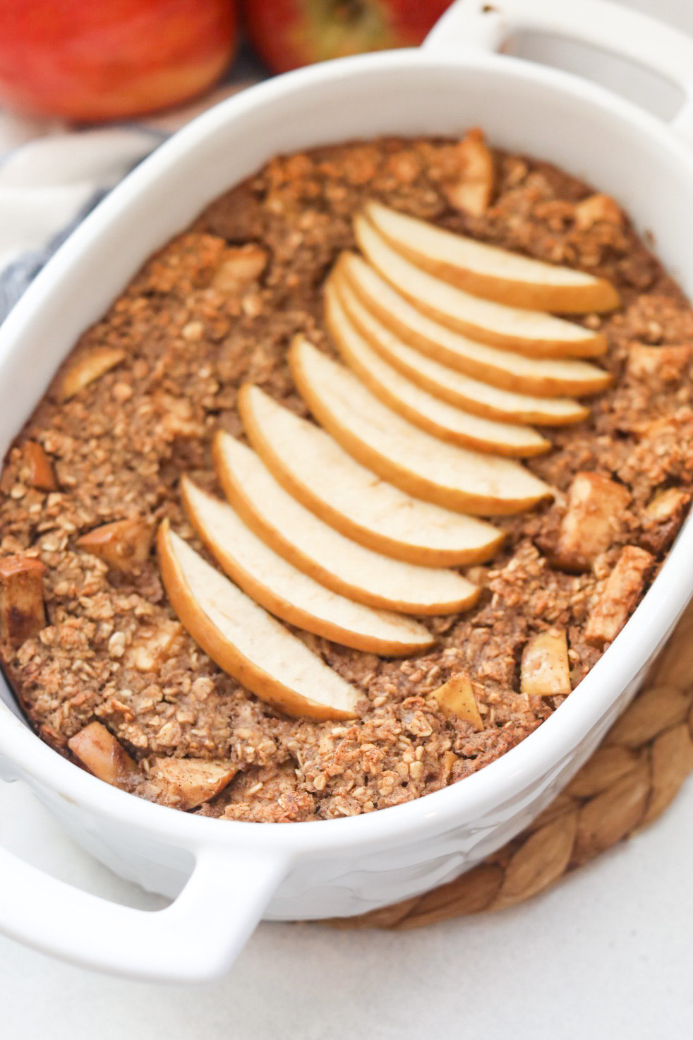 White oval baking dish with baked apple pie oatmeal topped with thin sliced red apples feathered on top. Two apples are out of focus in the top left corner of image.