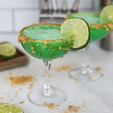 Two martini glasses with graham cracker crust rim, filled with Key Lime Pie Martini. Sliced limes beside the glass for styling purposes. Martini Kit in the background out of focus.