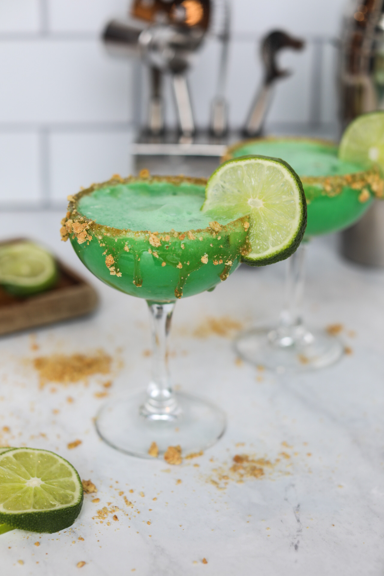 Two martini glasses with graham cracker crust rim, filled with Key Lime Pie Martini. Sliced limes beside the glass for styling purposes. Martini Kit in the background out of focus.