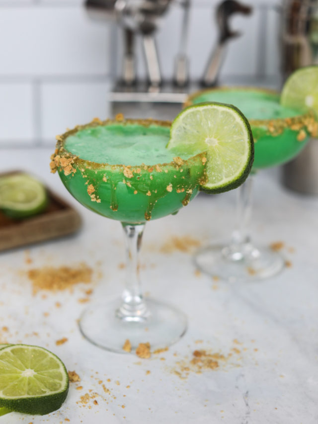 Key lime martini served in two cocktail glasses served with a graham cracker crust. Graham cracker crust and sliced limes added for styling purposes.