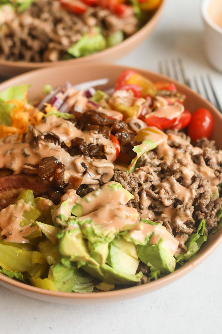 Bacon Cheeseburger Salad recipe image in a tan/brown bowl. Bacon Cheeseburger salad is filled with lettuce, tomatoes, onion, caramelized onions, cheddar cheese, avocado topped with a burger sauce. Another tan bowl is filled with the same ingredients for styling purposes.