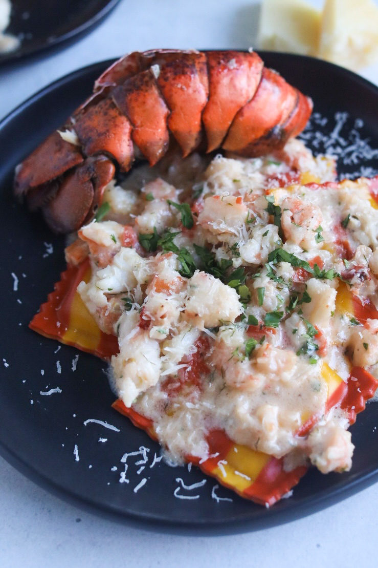 Lobster and shrimp ravioli completed dish on a black plate with Trader Joe's lobster ravioli that's striped in color with bright orange and yellow colors. Out of focus is a bright red lobster shell and a wedge of fresh parmesan cheese for styling purposes.