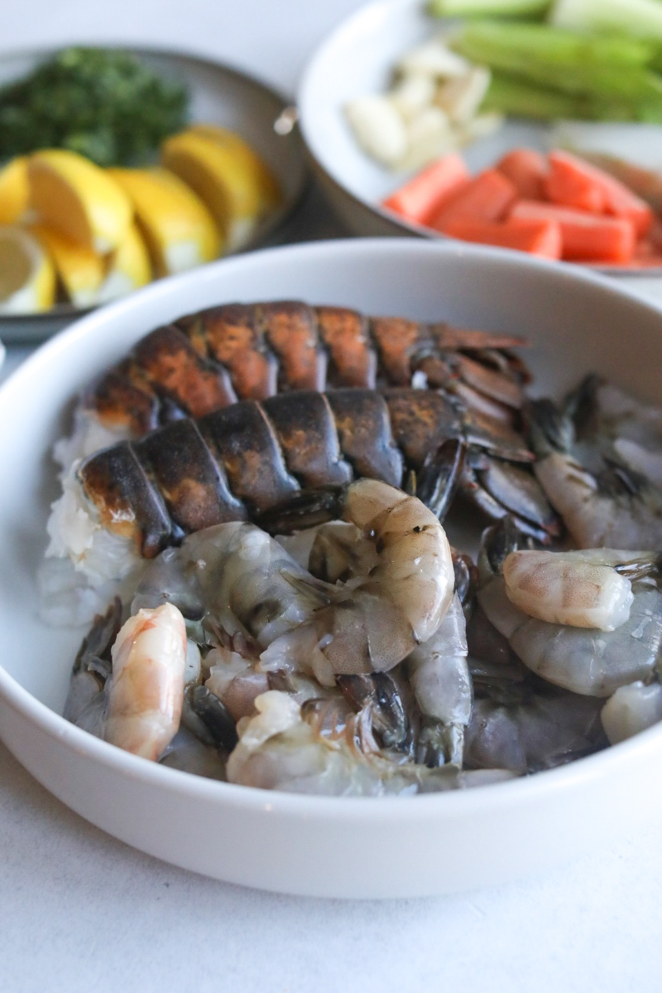 Two white bowls with seafood and seafood stock ingredients. The bowl in the center has fresh shelled shrimp and two lobster tails. The white bowl to the right has carrots, celery and onion that's out of focus. The left plate is gray and has lemon wedges and fresh herbs (dill and parsley).