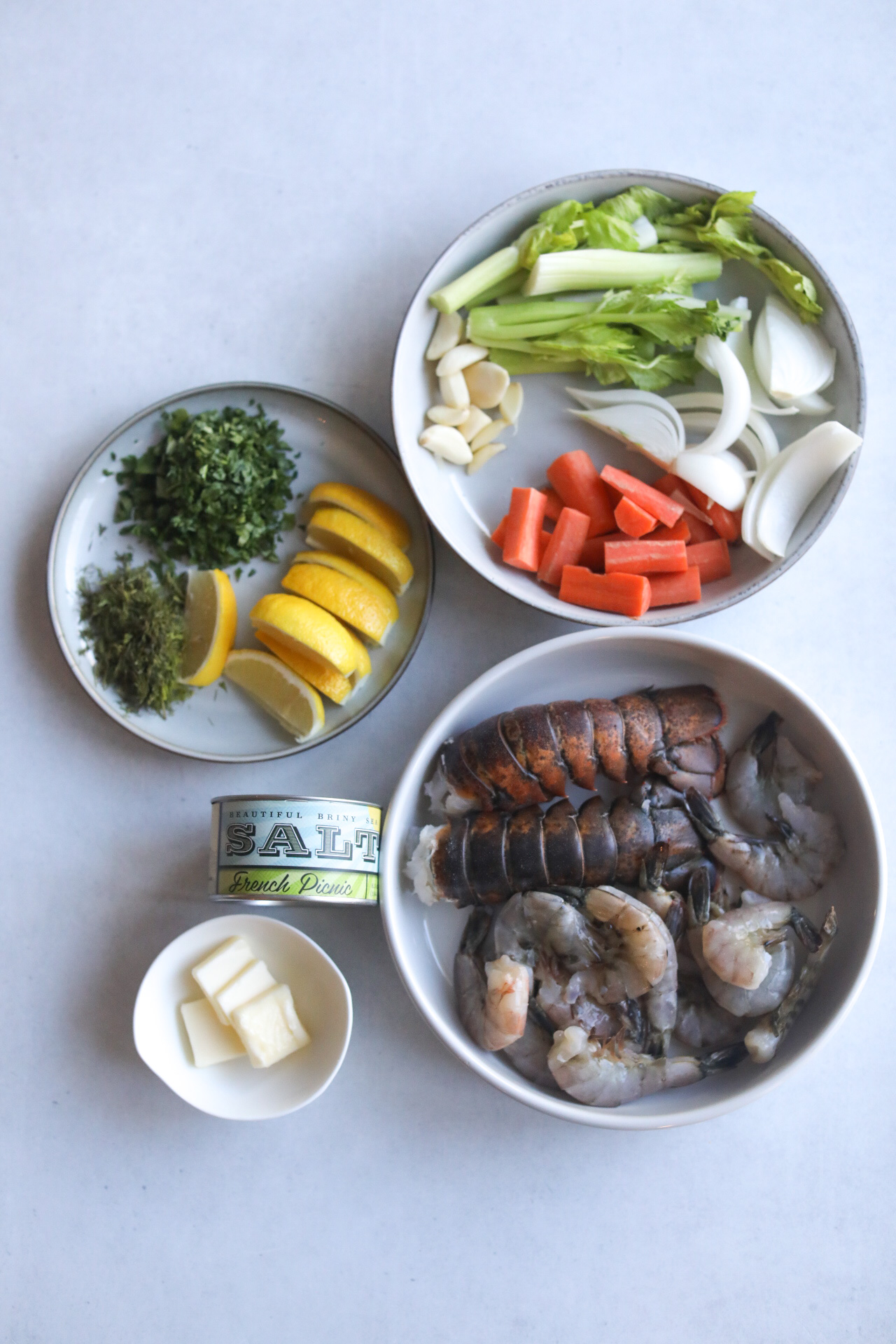 Lobster and Shrimp with Stock Ingredients with two bowls and one gray plate. On the right side the first bowl has raw shrimp and lobster tails in their shells. The top right bowl and seafood stock ingredients (carrots, onion, garlic and celery). The left gray plate has lemon wedges and fresh herbs chopped (parsley and dill). The bottom white small bowl has butter slices. A seasoning blend can from Beautiful Briny Sea with a seasoning label of French Picnic on the front.