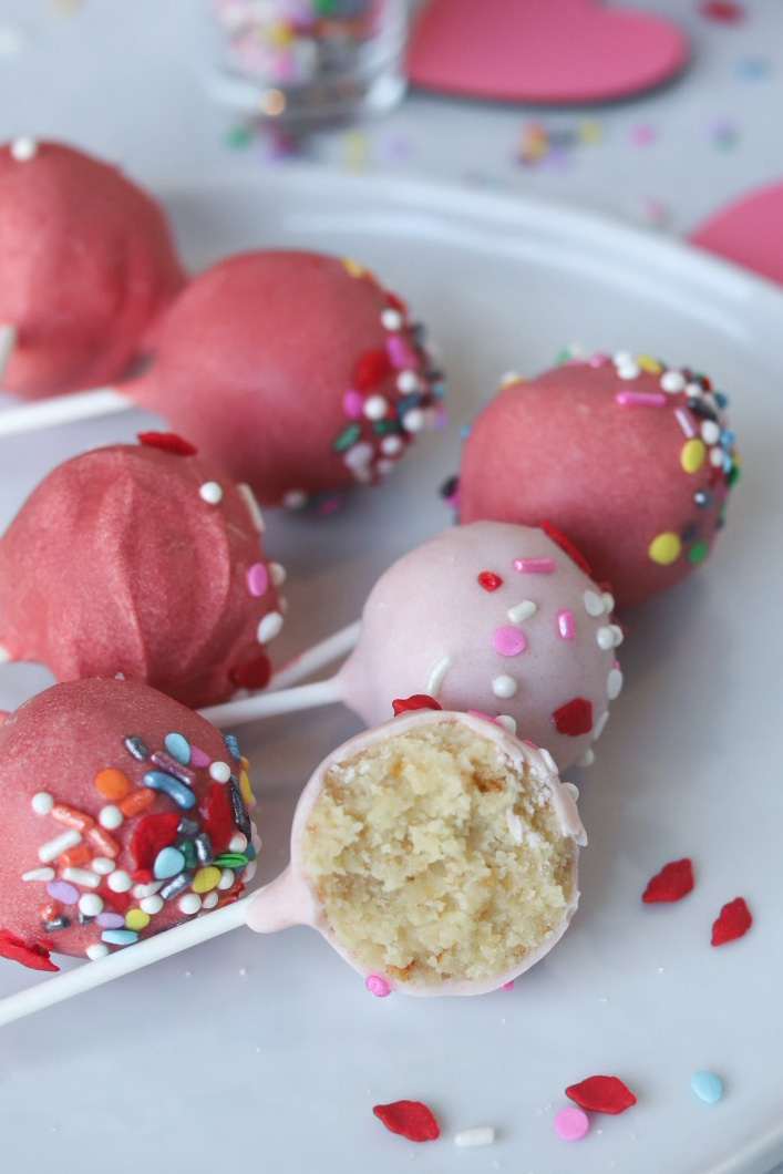 Valentine's themed cake pops in light and darker shades or pink. Six cake pops lying flat with a cake pop cut in half sitting in front of cake pops with sprinkles. The bottom of a clear shot glass filled with sprinkles out of focus behind the cake pops as well as glitter wooden hearts in dark pink color for styling purposes.