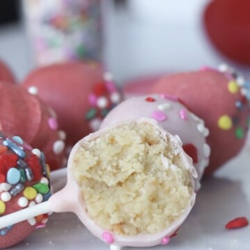 Valentine's themed cake pops in light and darker shades of pink. Three cake pops lying flat with a cake pop cut in half sitting in front of cake pops with sprinkles. A clear shot glass filled with sprinkles out of focus behind the cake pops as well as glitter wooden hearts in dark pink color for styling purposes.