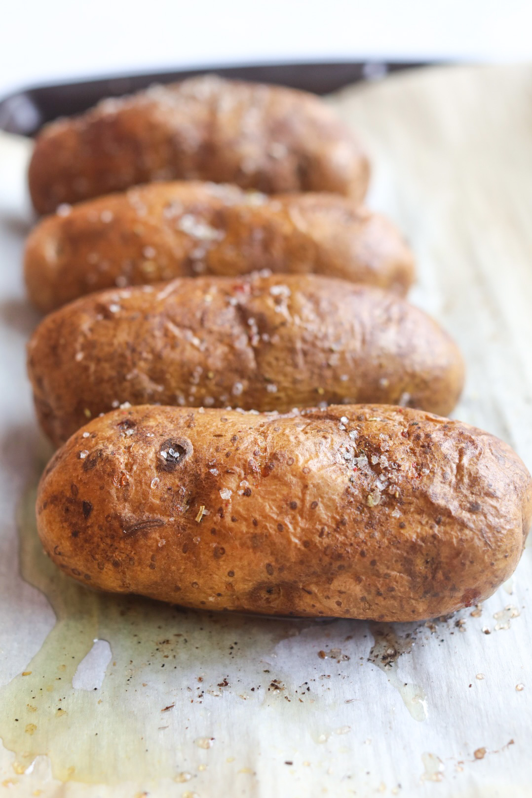 Four baked potatoes covered with oil and coarse salt. Potatoes are baked and on a lined baking sheet with parchment paper.