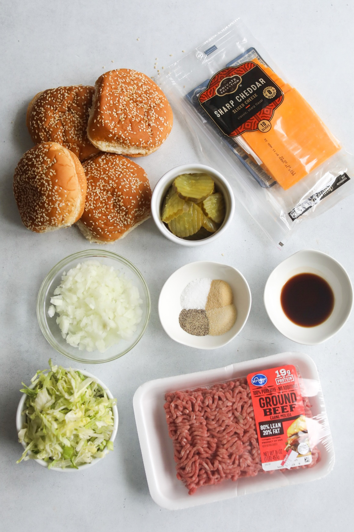 Big Mac Sloppy Joe ingredients, flat lay style. Ground beef packaged, bowl of shredded lettuce, cup of diced white onion, four sesame seed buns, cheddar cheese slices, cup of pickles, burger seasonings and W sauce.