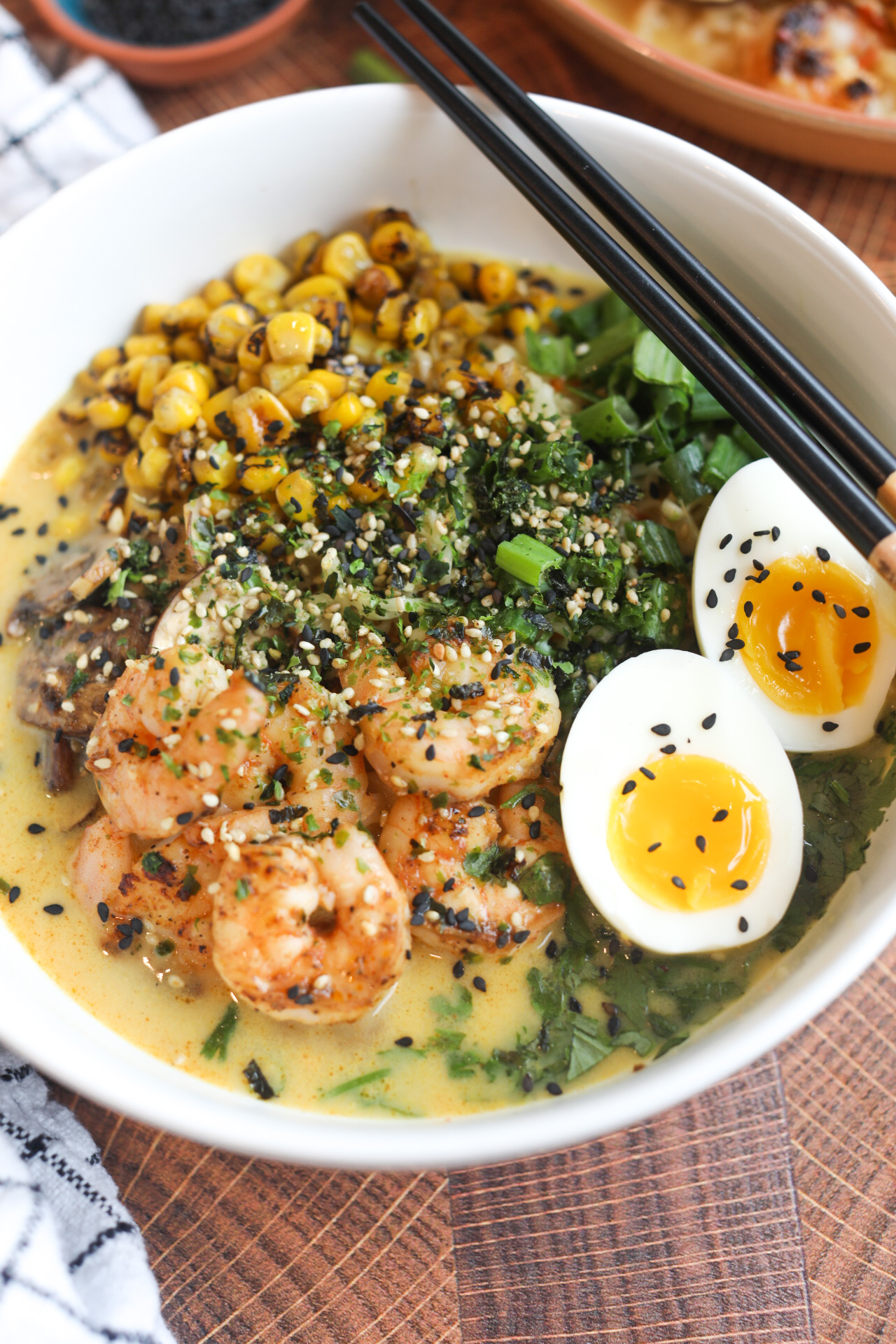 White bowl with shrimp ramen noodles topped with soft boiled egg, cilantro, parsley, corn and mushrooms. Ramen bowl is topped with sesame seeds and chopsticks and added to the right of the bowl for styling purposes. The dish has a black and white checkered towel and an additional bowl in the top right corner of the pic for added dimension.