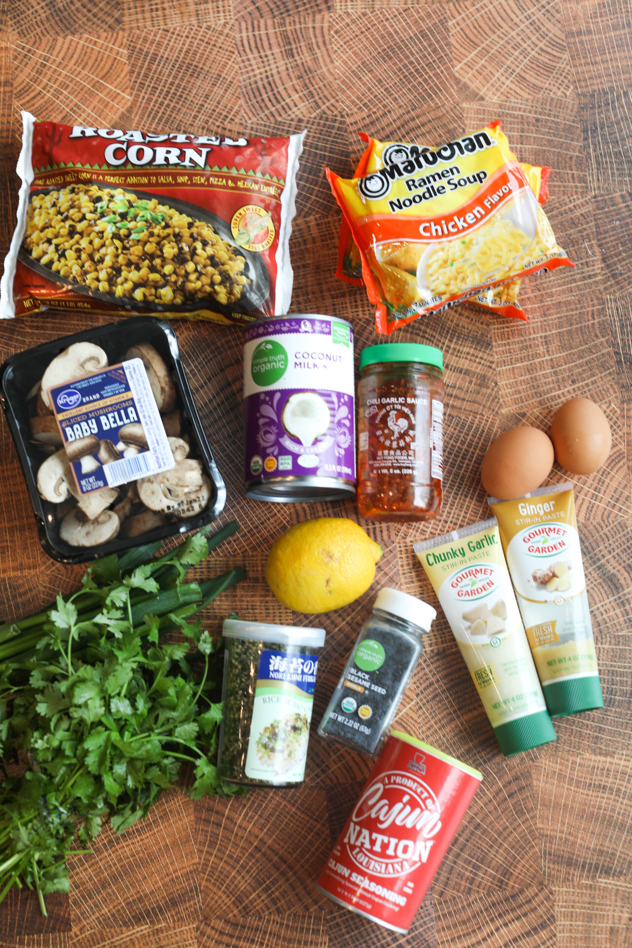 Shrimp Ramen Noodles Bowl ingredients. Ingredients are laid in a flay lay style in a box. From left to right; bag of roasted corn, two packages or ramen noodles, box of bella mushrooms, can of coconut milk, jar of garlic chili paste, two brown eggs, bunch of cilantro, lemon, can of furikake, can of sesame seeds, can of cajun seasoning, tubes of garlic and ginger paste.