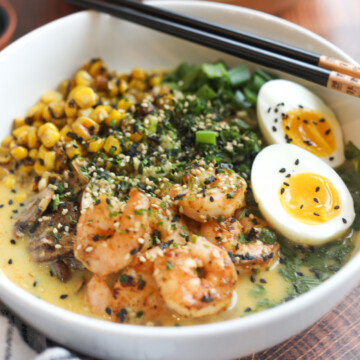 White bowl with shrimp ramen noodles topped with soft boiled egg, cilantro, parsley, corn and mushrooms. Ramen bowl is topped with sesame seeds and chopsticks and added to the right of the bowl for styling purposes. The dish has a black and white checkered towel and an additional bowl in the top right corner of the pic for added dimension.