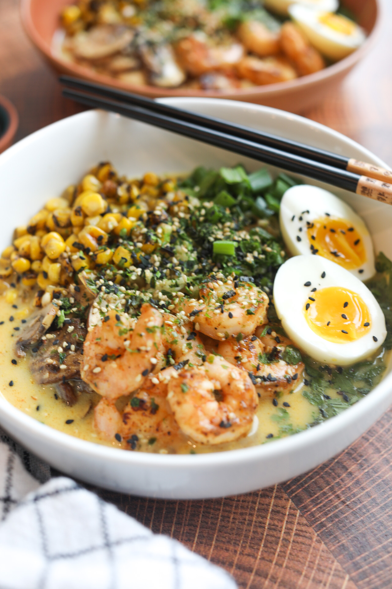 White bowl with shrimp ramen noodles topped with soft boiled egg, cilantro, parsley, corn and mushrooms. Ramen bowl is topped with sesame seeds and chopsticks and added to the right of the bowl for styling purposes. The dish has a black and white checkered towel and an additional bowl in the top right corner of the pic for added dimension.