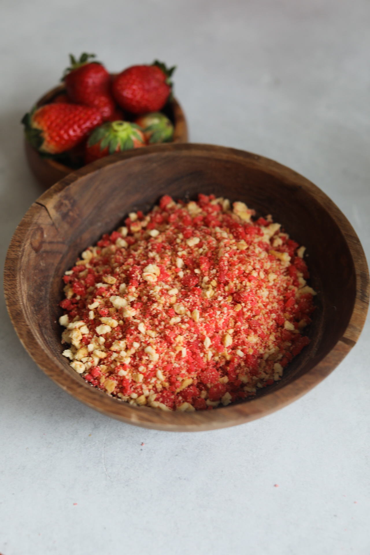 Strawberry cake pop crunch topping in a small wooden bowl with a small bowl of fresh strawberries for styling purposes.