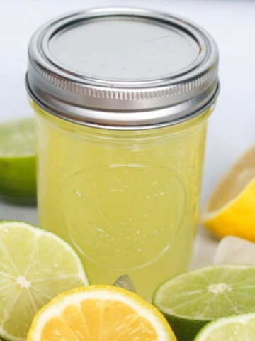 Sour mix in a short glass mason jar with metal lid. Fresh lemons and limes cut in half surrounding glass for styling purposes.