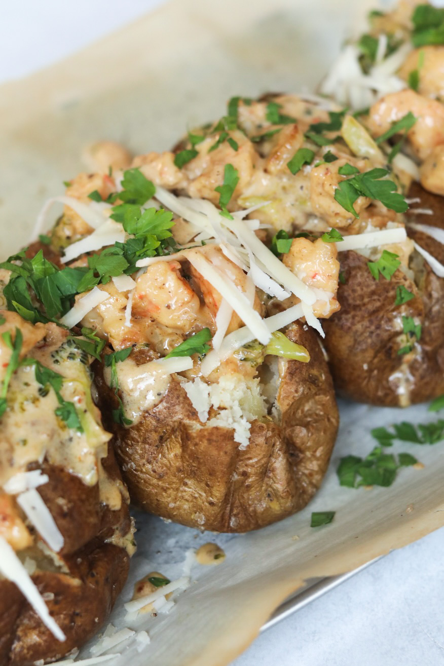 Four baked potatoes baked and topped with Cajun shrimp cream sauce. Baked potatoes are topped with additional shredded cheese and fresh parsley.