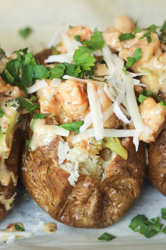 Three baked potatoes, one focused in the center of the image, baked and topped with Cajun shrimp cream sauce. Baked potatoes are topped with additional shredded cheese and fresh parsley.