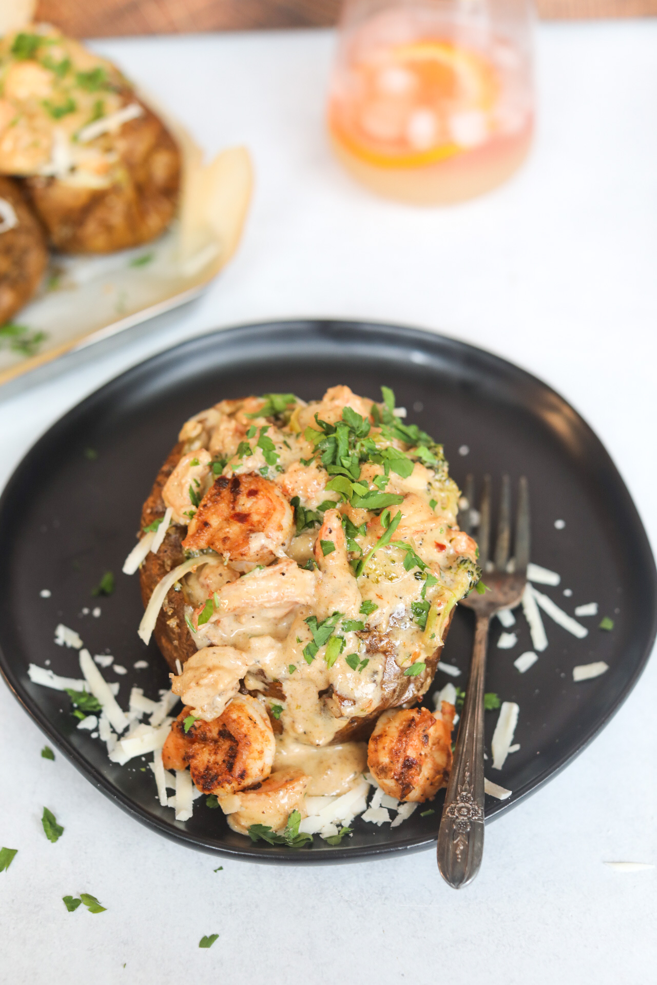 Loaded Shrimp Baked Potato on a black dining plate with extra cream sauce, shrimp and shredded cheese around image for styling purposes. The image of the baked potato is slightly zoomed in to solely focus on one potato. In the background out of focus is one glass filled with an orange cocktail and additional baked potatoes with cream sauces added for styling purposes.