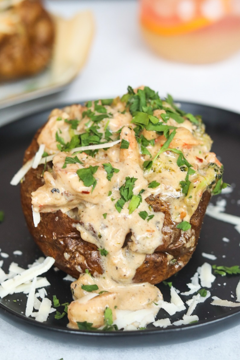 Loaded Shrimp Baked Potato on a black dining plate with extra cream sauce and shredded cheese around image for styling purposes. The image of the baked potato is zoomed in to solely focus on one potato.