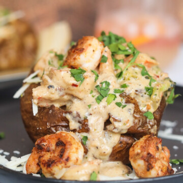 Loaded Shrimp Baked Potato on a black dining plate with extra shrimp in cream sauce and shredded cheese around image for styling purposes. The image of the baked potato is zoomed in to solely focus on one potato.
