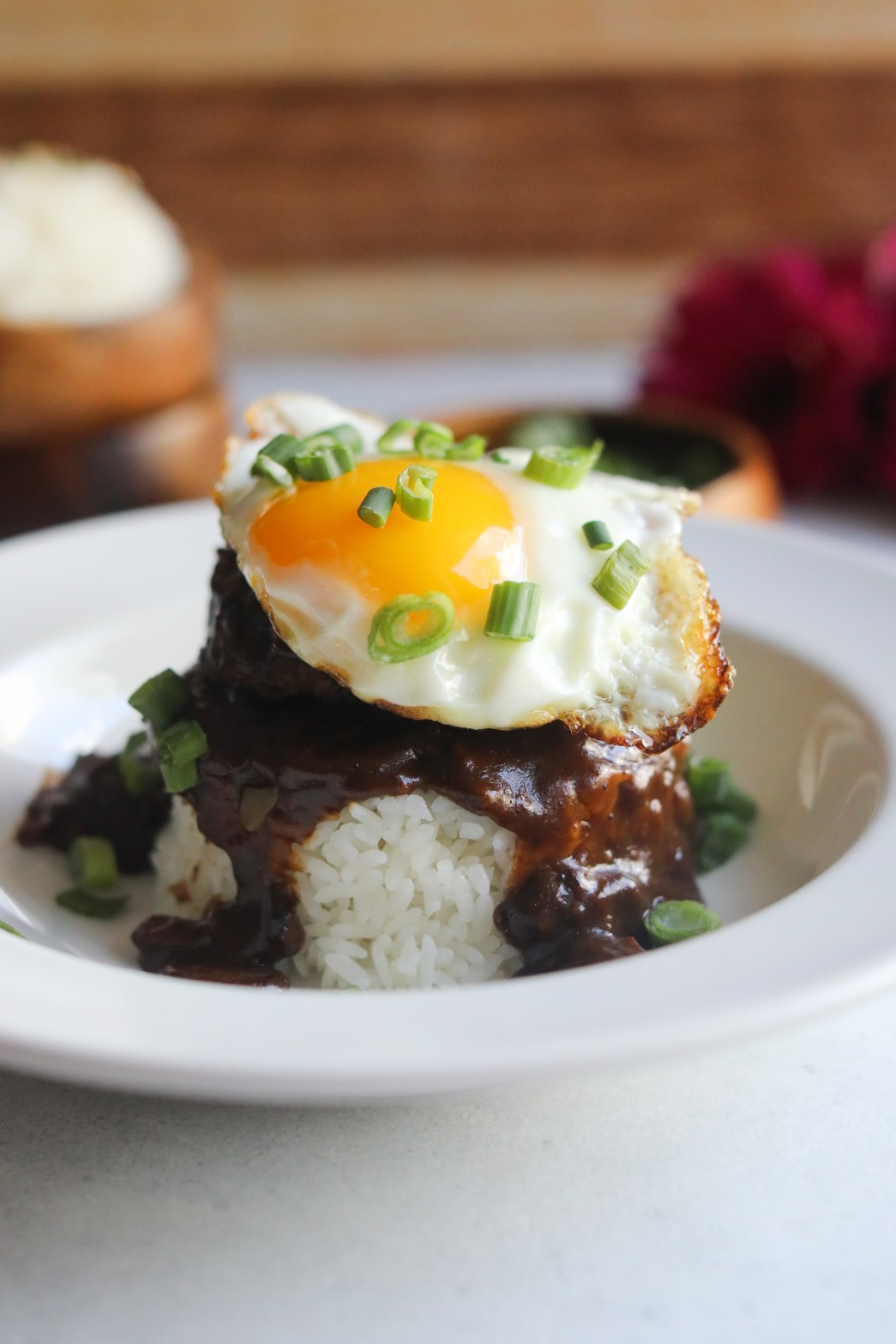 Loco Moco Recipe in a white shallow bowl. A mound of rice in an oval/circular shape is topped with brown gravy, hamburger patty and topped with a sunny side up egg. Dish is topped with chopped green onions and in the background out of focus are two wooden bowls stacked with extra white rice (on the left) and on the right images of dark pink flowers.