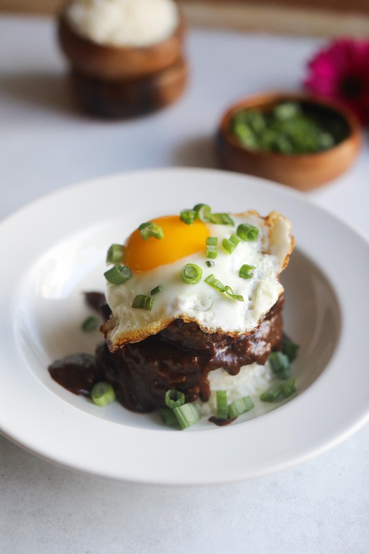 Loco Moco Recipe in a white shallow bowl. A mound of rice in an oval/circular shape is topped with brown gravy, hamburger patty and topped with a sunny side up egg. Dish is topped with chopped green onions and in the background out of focus are two wooden bowls stacked with extra white rice (on the left) and on the right images of dark pink flowers.