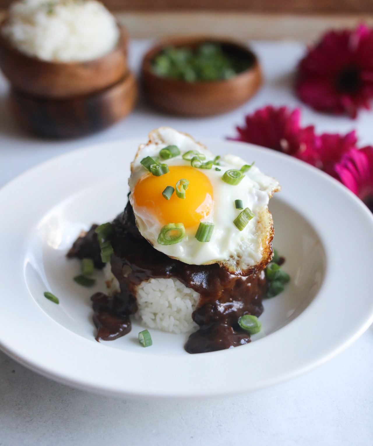 Loco Moco Recipe in a white shallow bowl. A mound of rice in an oval/circular shape is topped with brown gravy, hamburger patty and topped with a sunny side up egg. Dish is topped with chopped green onions and in the background out of focus are two wooden bowls stacked with extra white rice (on the left) and on the right images of dark pink flowers.