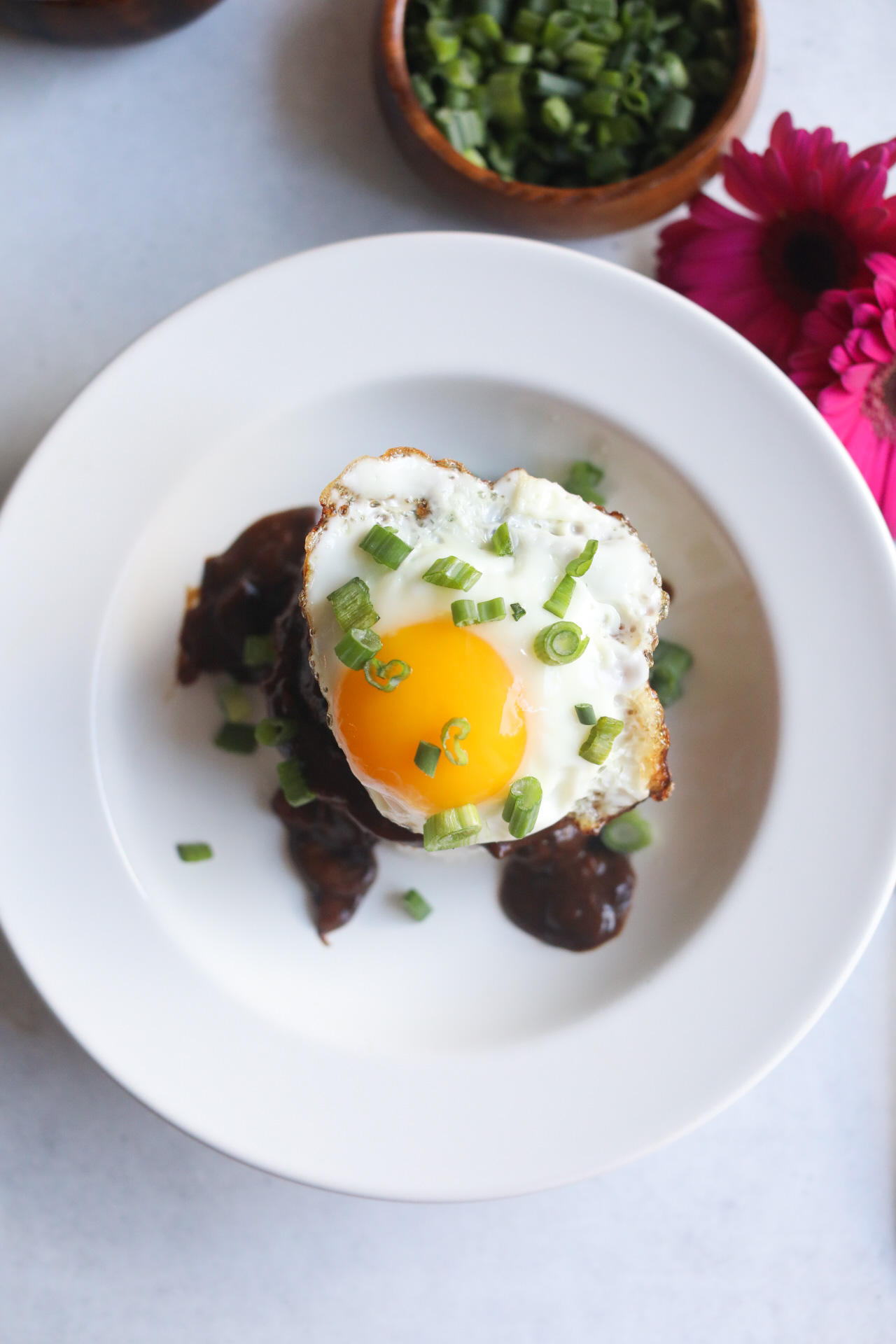 Loco Moco Recipe in a white shallow bowl with top down angle viewing the image from "above". Dark pink flowers and a wooden bowl with chopped green onions added for styling purposes.