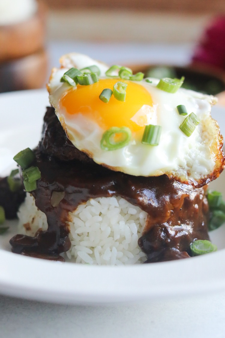 Loco Moco Recipe in a white shallow bowl. A mound of rice in an oval/circular shape is topped with brown gravy, hamburger patty and topped with a sunny side up egg. Dish is topped with chopped green onions and in the background out of focus are two wooden bowls stacked with extra white rice (on the left) and on the right images of dark pink flowers.