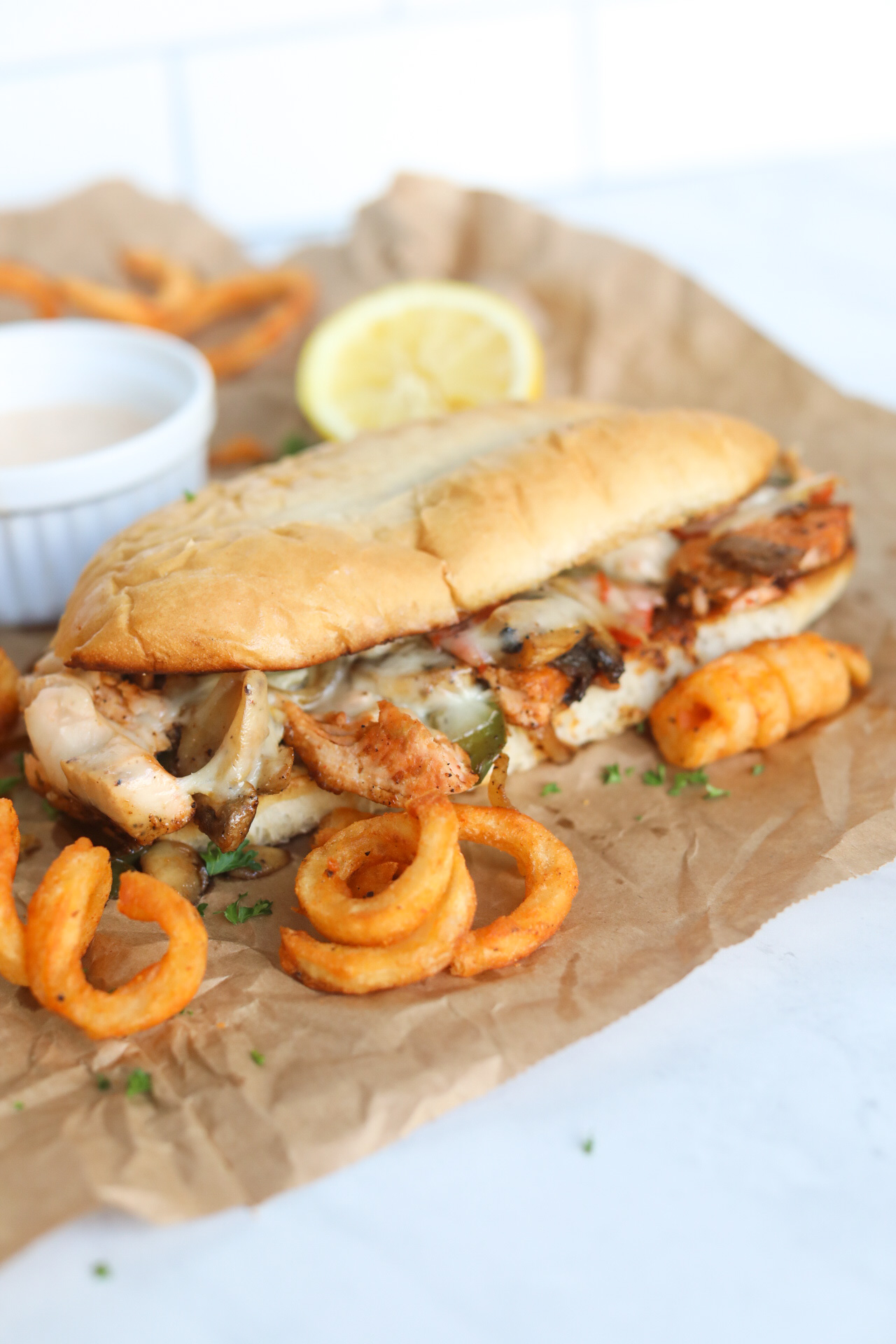 Salmon Cheesesteak with remoulade sauce in a white ramekin. Curly fries and a half of a used lemon is added for styling purposes. 