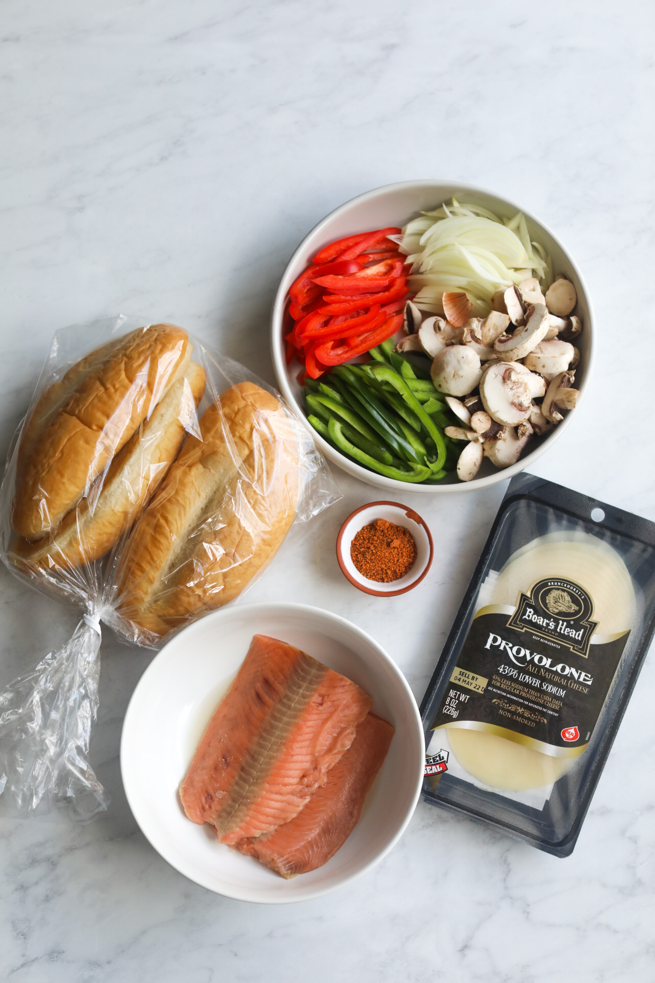 Salmon Cheesesteak ingredients. Hoagie rolls in a clear bag, grey bowl with red and green peppers, onions and mushrooms. Small bowl of Cajun seasoning. Plate of two pieces of salmon and a package of provolone cheese. 