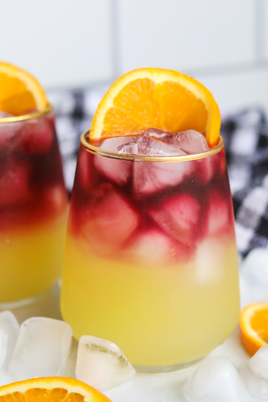 Two Sangria Margarita in a stemless wine glass rimmed in gold. Ice cubes, and a sliced orange on the side of the glass is added for styling purposes. Ice cubes, black and white towel and additional sliced oranges, lemon and limes are added for styling purposes.