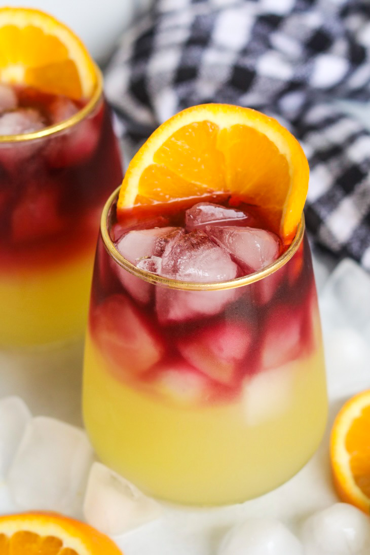 One sangria margarita in center of image with one additional, identical drink behind the cocktail scaled off the image. Black and white kitchen towel and sliced oranges and ice cubes added for styling purposes.