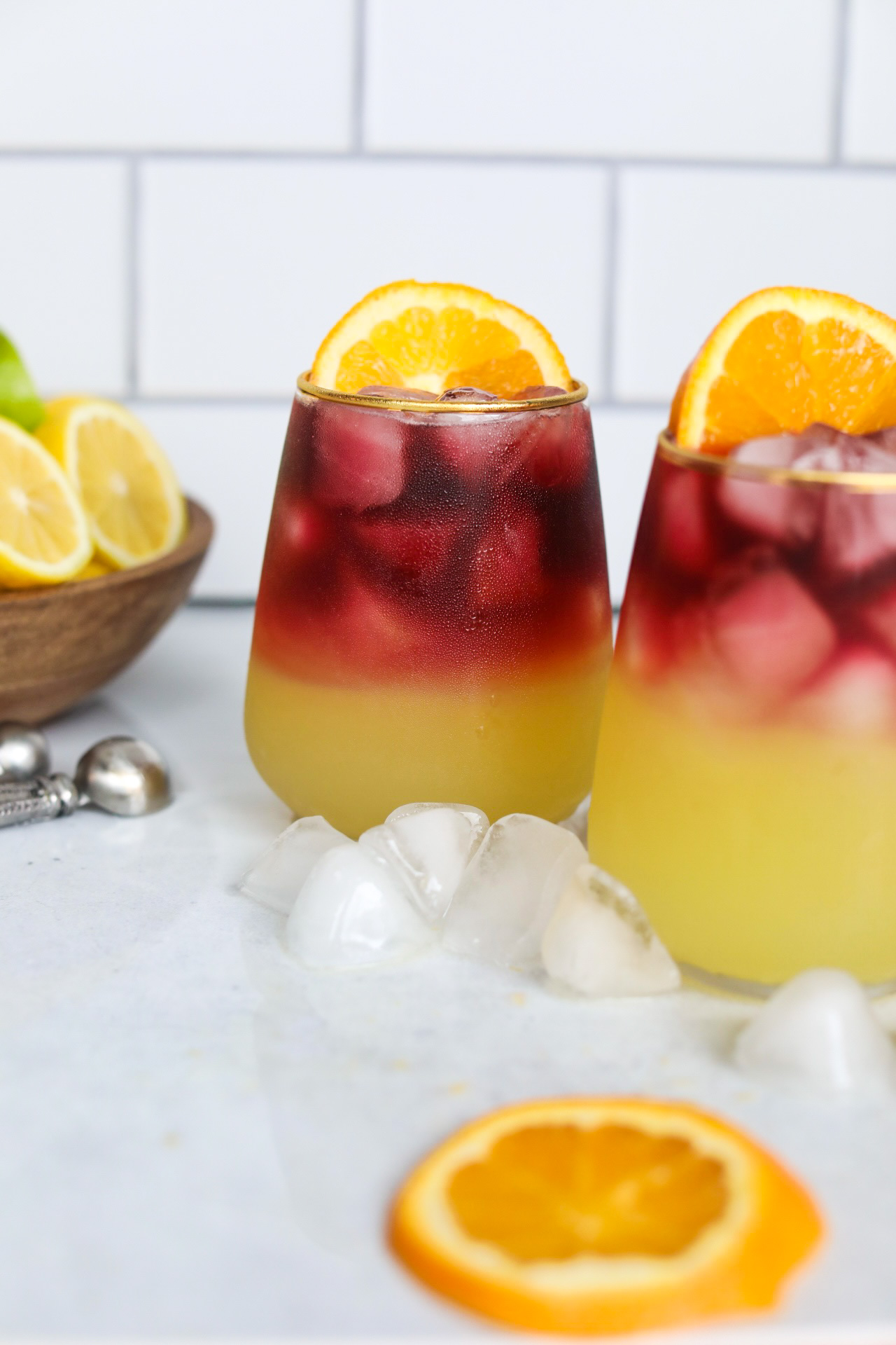 Two Sangria Margarita in a stemless wine glass rimmed in gold. Ice cubes, and a sliced orange on the side of the glass is added for styling purposes. Ice cubes, black and white towel and additional sliced oranges, lemon and limes are added for styling purposes.