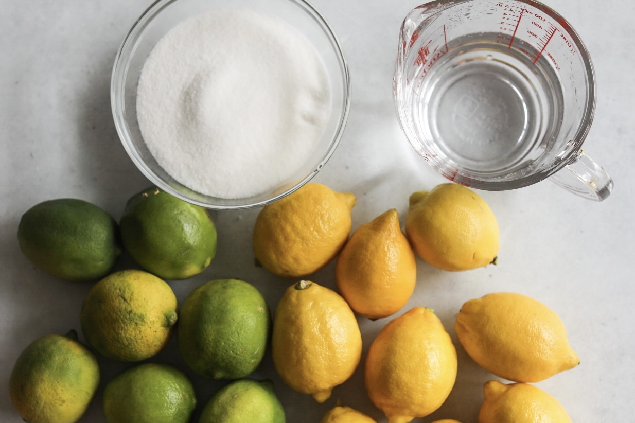 Whole lemons and limes bunched together on the bottom of the image with a bowl of white sugar and a glass measuring cup filled with water.