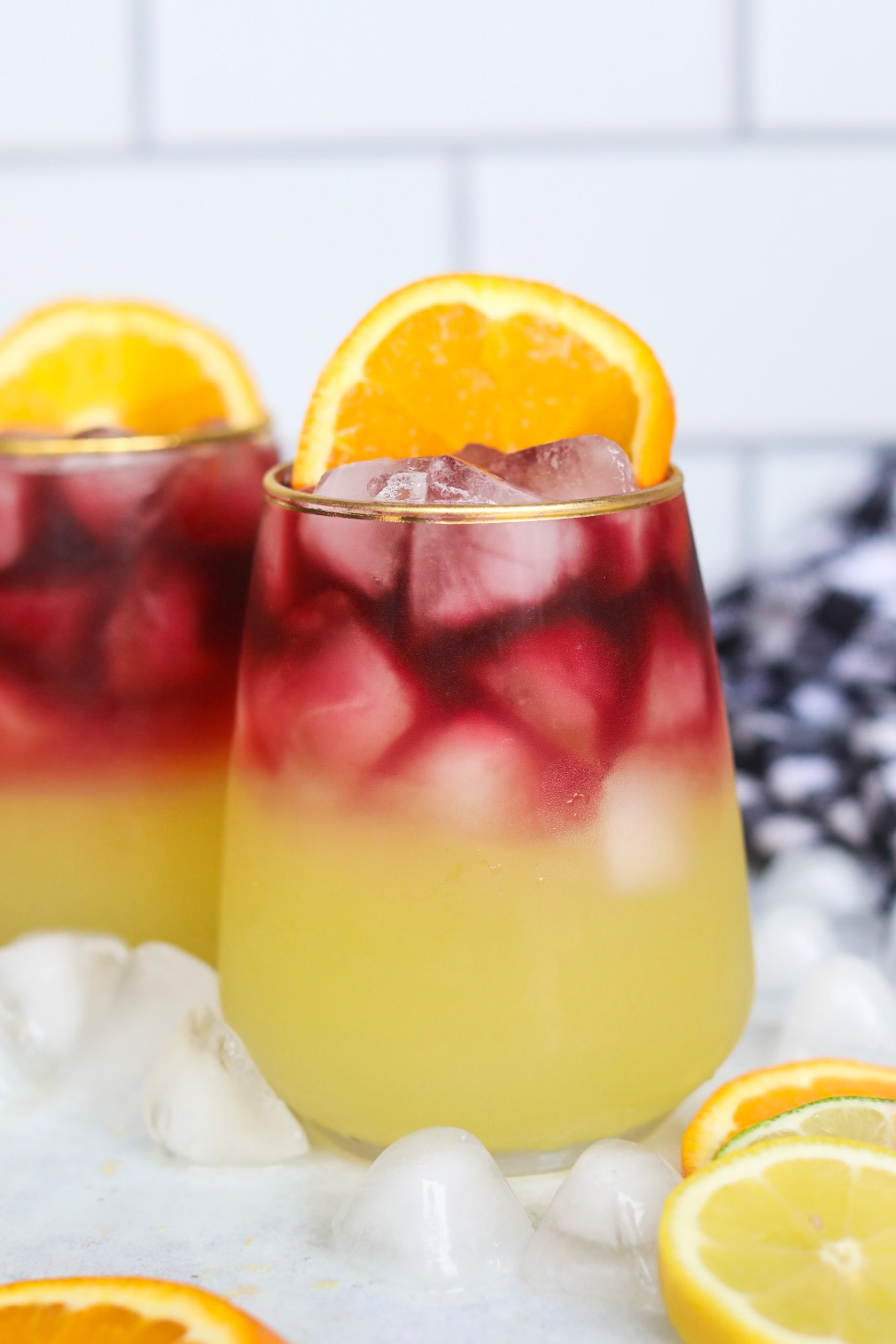 Two Sangria Margarita in a stemless wine glass rimmed in gold. Ice cubes, and a sliced orange on the side of the glass is added for styling purposes. Ice cubes, black and white towel and additional sliced oranges, lemon and limes are added for styling purposes.