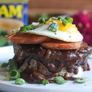 Spam Loco Moco Recipe plated on a white flat plate. Bed of sticky rice layered with brown gravy topped with two slices of spam topped with a sunny side up egg. Dark pink flowers, wooden bowls with sticky rice added for styling purposes as well as an opened can of Spam.