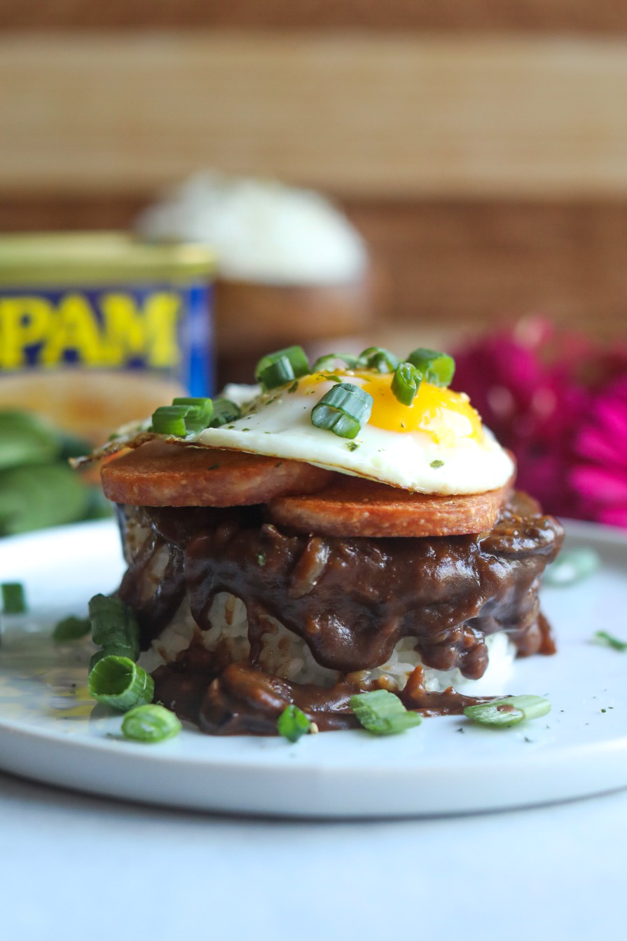 Spam Loco Moco Recipe plated on a white flat plate. Bed of sticky rice layered with brown gravy topped with two slices of spam topped with a sunny side up egg. Dark pink flowers, wooden bowls with sticky rice added for styling purposes as well as an opened can of Spam.