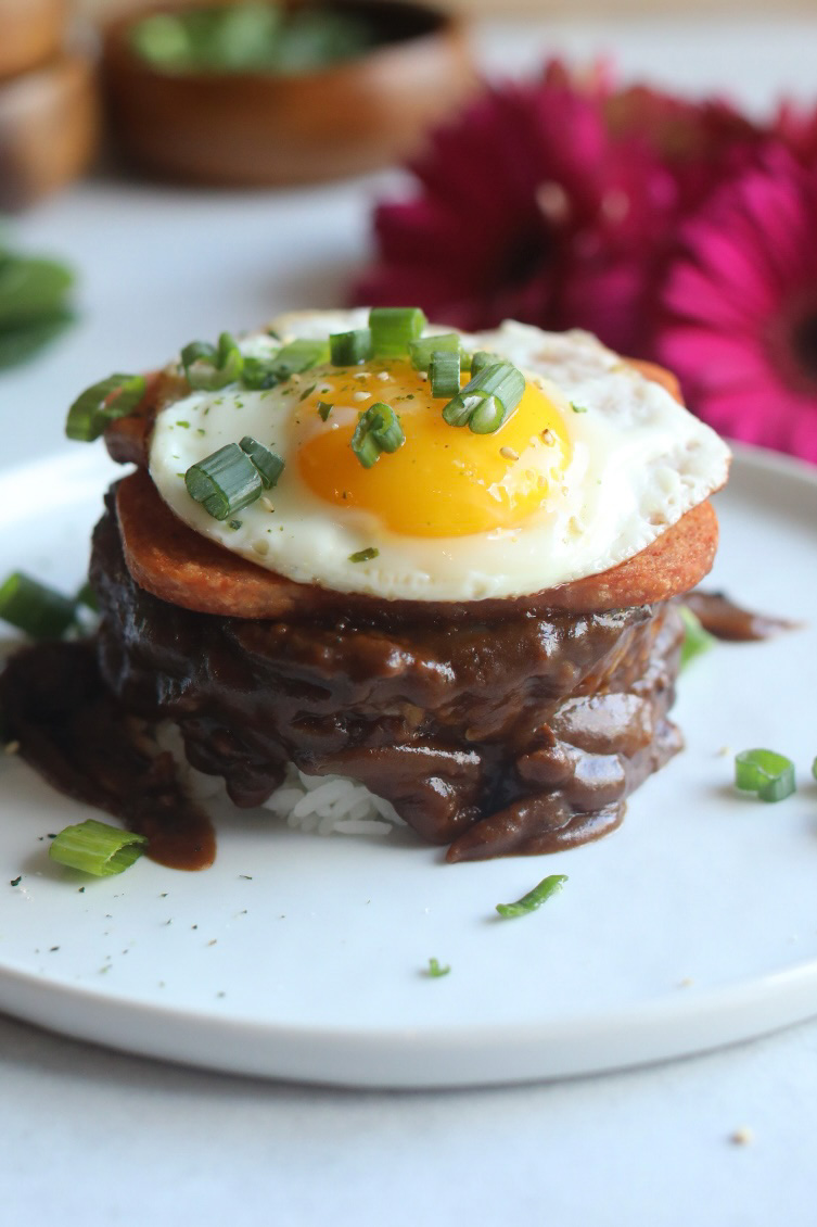 Spam Loco Moco Recipe plated on a white flat plate. Bed of sticky rice layered with brown gravy topped with two slices of spam topped with a sunny side up egg. Dark pink flowers and wooden bowls with chopped green onions added for styling purposes.