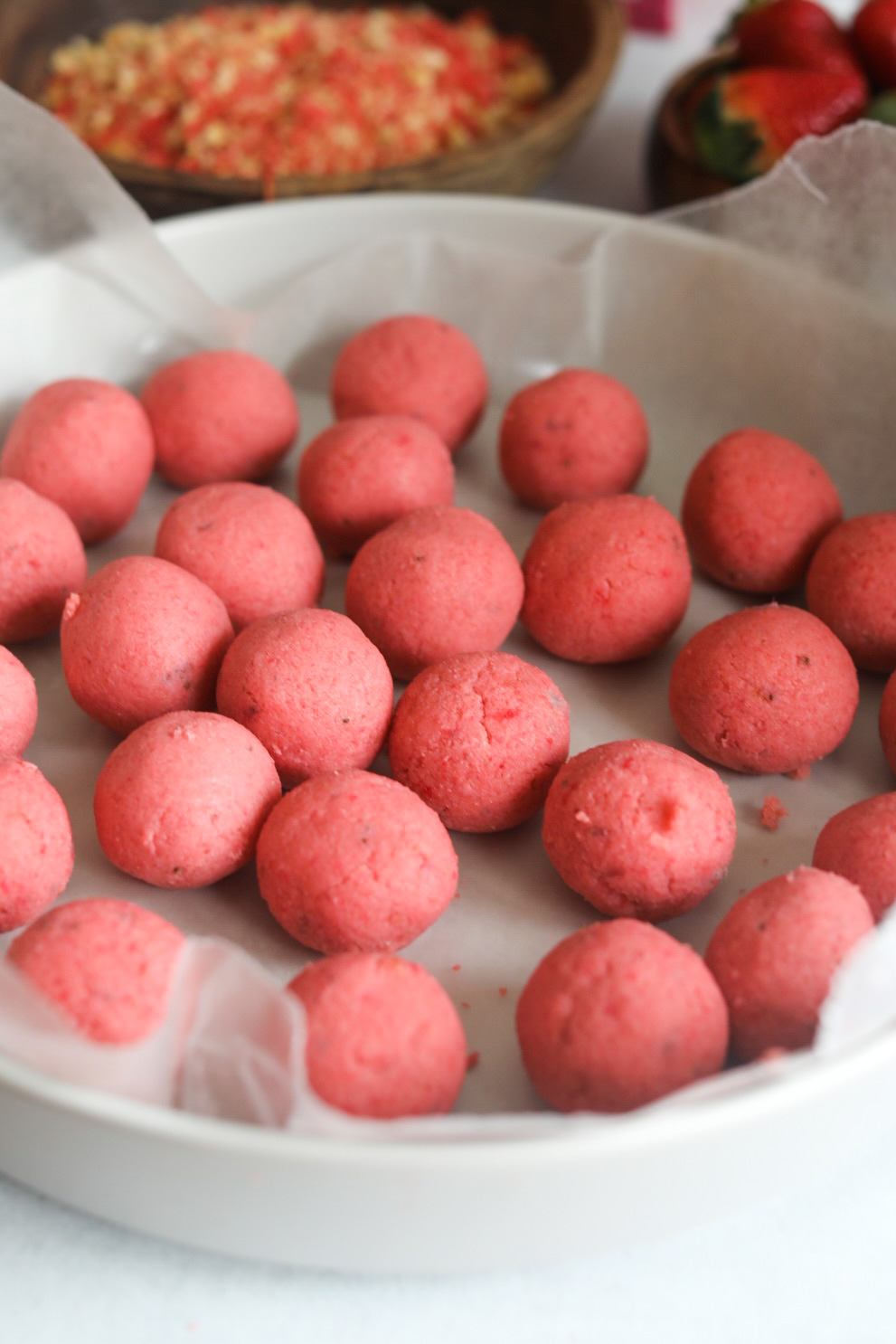 Strawberry cake pop in balls, about 24 balls in a lined grey large deep dish. Out of focus is the strawberry crunch in a wooden bowl.