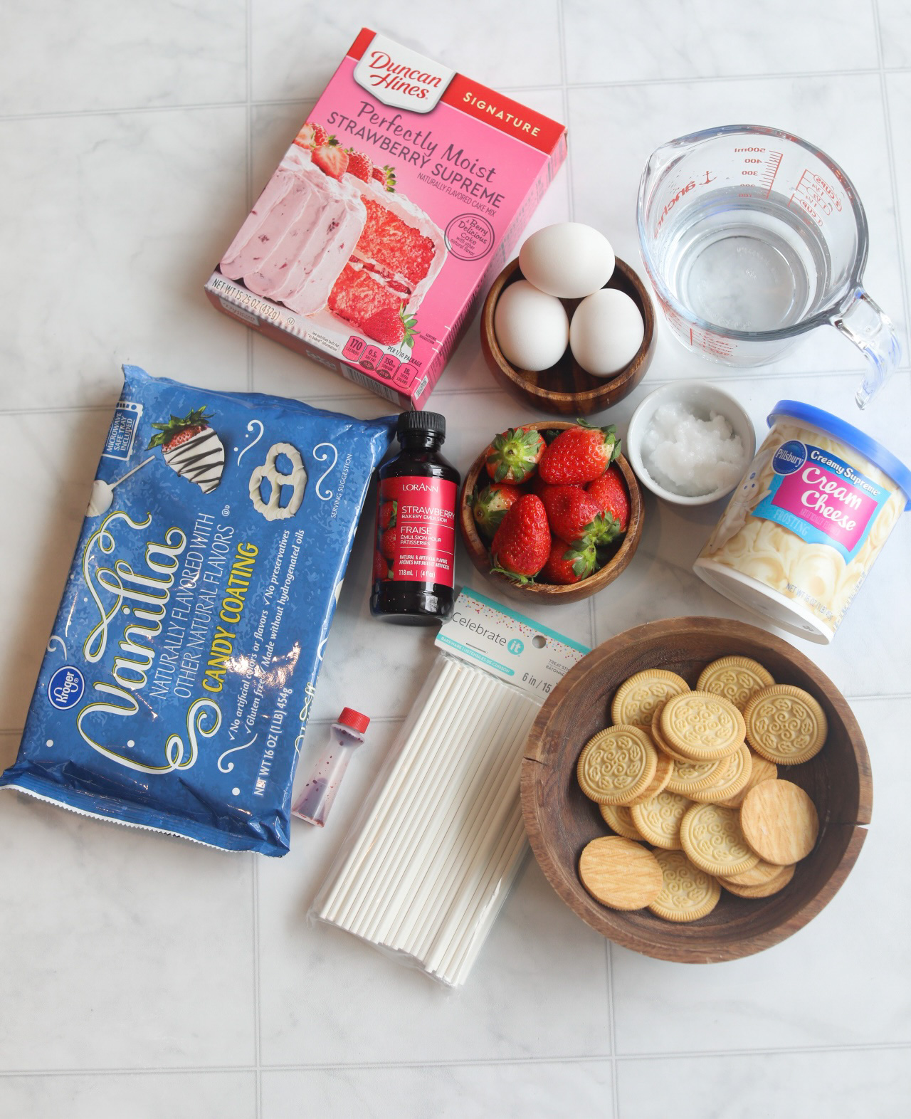 Strawberry cake pops with crunch topping ingredients. From left to right, strawberry boxed cake mix, three eggs in a wooden bowl, clear measuring glass filled with water. small white bowl filled with coconut oil, cream cheese frosting in blue and white container, wooden bowl with vanilla cookies, cake pop sticks in clear bag, white cholate bark in a blue bag, wooden bowl filled with whole-fresh strawberries and small bottle of strawberry emulsion.