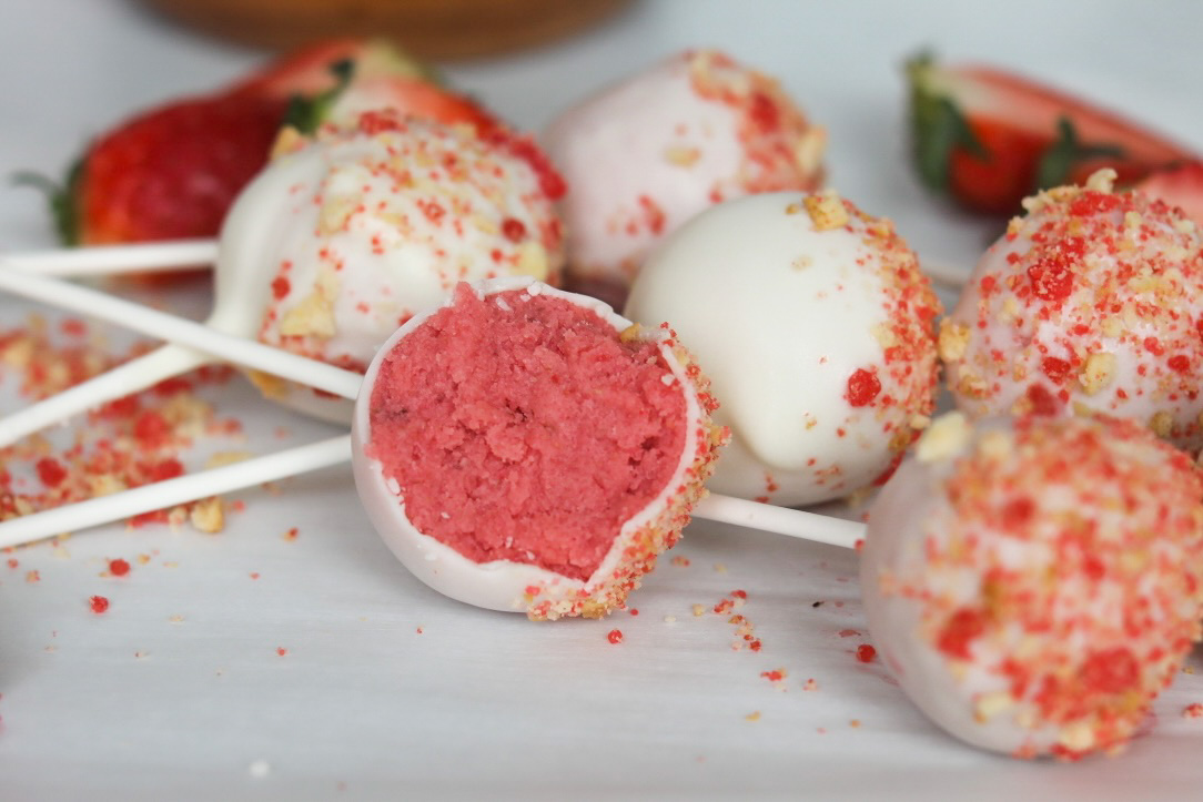 Close up image of Strawberry Cake Pops lying flat on white parchment paper. One cake pop has been bitten through the middle to show the exposed cake beneath the white chocolate covering.