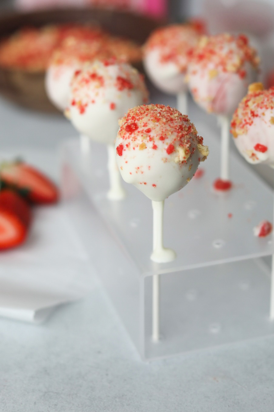 Six strawberry cake pops in a clear cake pop stand. One halved strawberry added for styling purposes on the right side of the cake pops. Two wooden bowls out of focus in the background added for styling purposes filled with strawberry crunch and fresh strawberries.