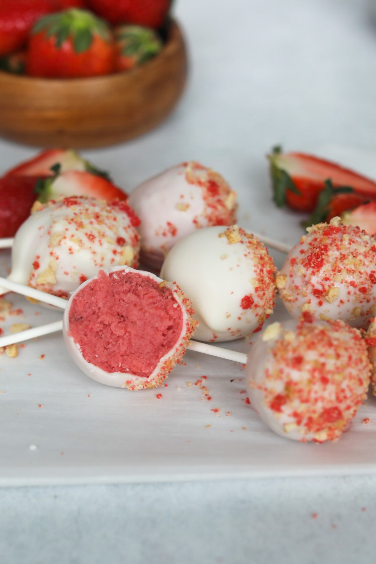 Image of Strawberry Cake Pops lying flat on white parchment paper. One cake pop has been bitten through the middle to show the exposed cake beneath the white chocolate covering.