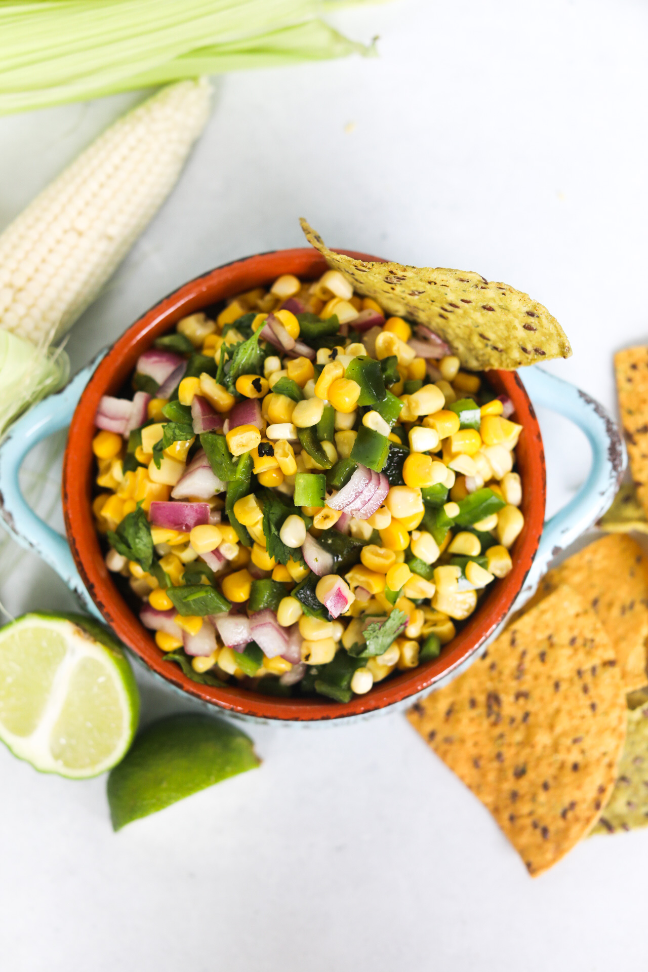 Corn salsa in a blue serving bowl in aerial view. Corn chip added for styling purposes as well as additional tortilla chips, sliced lime wedges and an ear of corn.