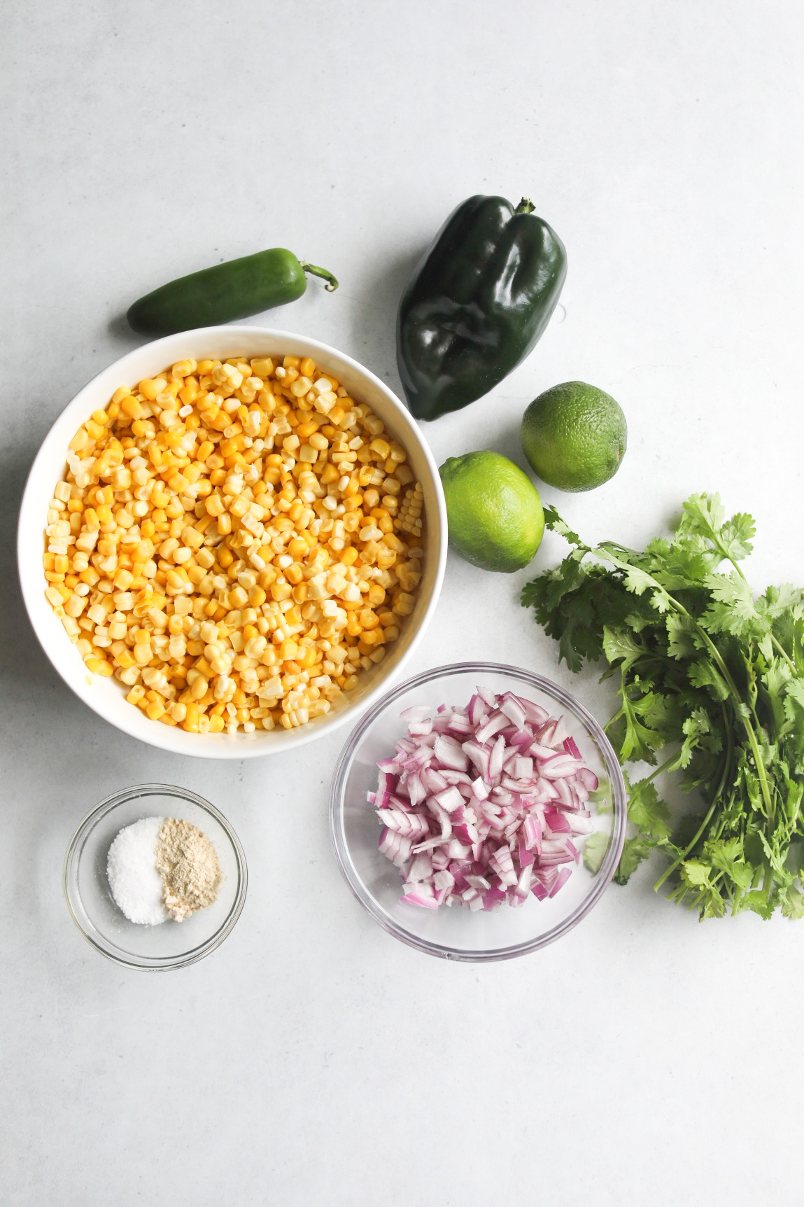 Chipotle Copycat Corn Salsa ingredients on a flat lay. Ingredients are a medium sized white bowl filled with corn. Jalapeno, poblano pepper, two limes, cilantro, glass bowl with diced red onion, small glass bowl with salt and garlic powder.