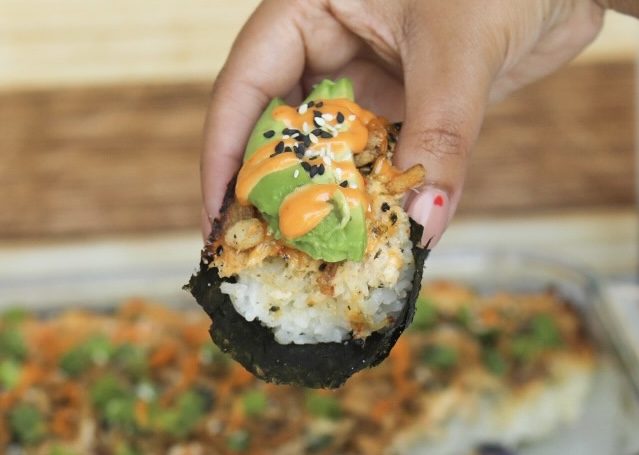 Hand holding baked sushi bake wrapped in seaweed sheets. Sushi bake is topped with sliced avocado, spicy mayo and sesame seeds.