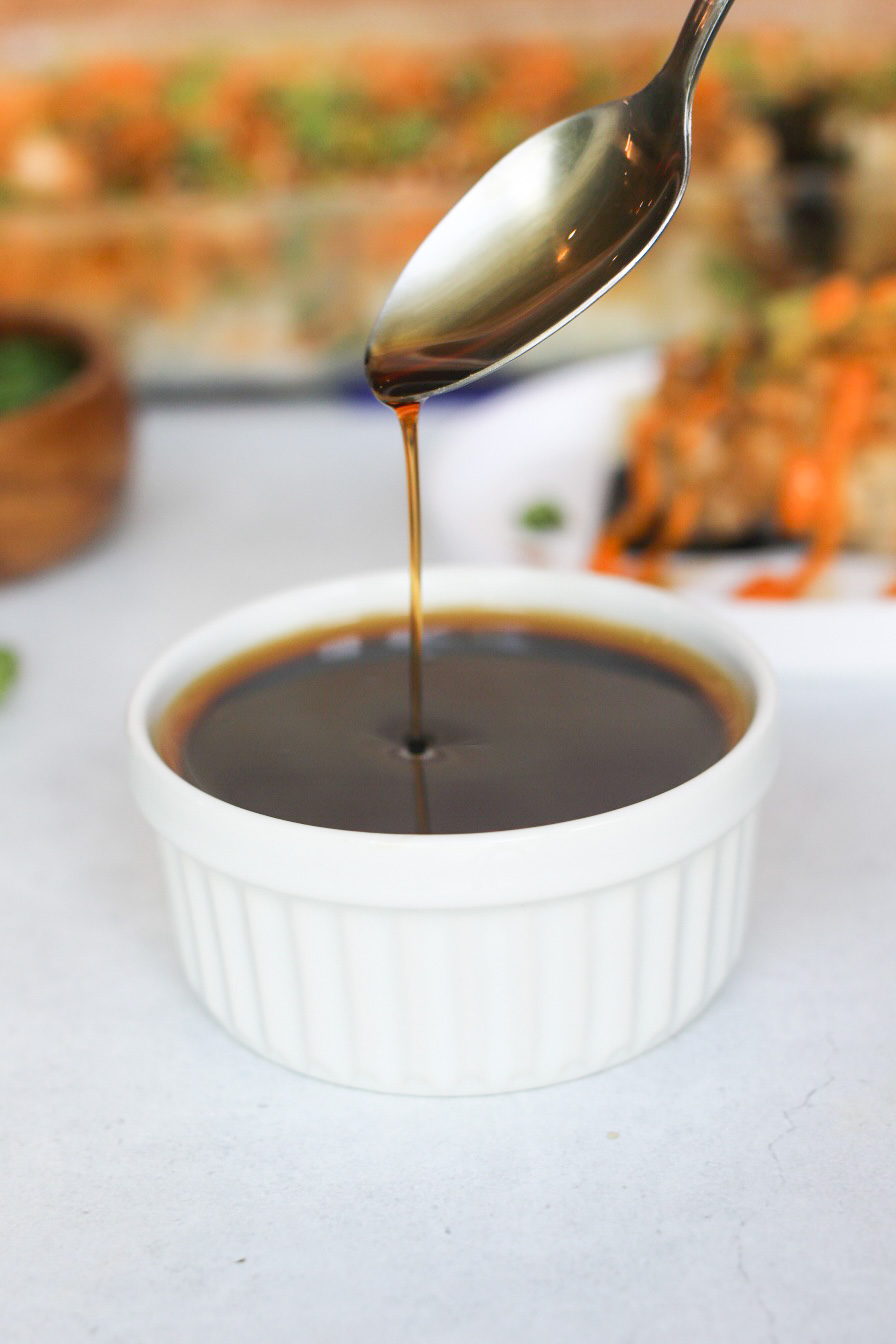 Eel sauce in a white ramekin. Spoon is ladled over the white ramekin pouring eel sauce into cup.