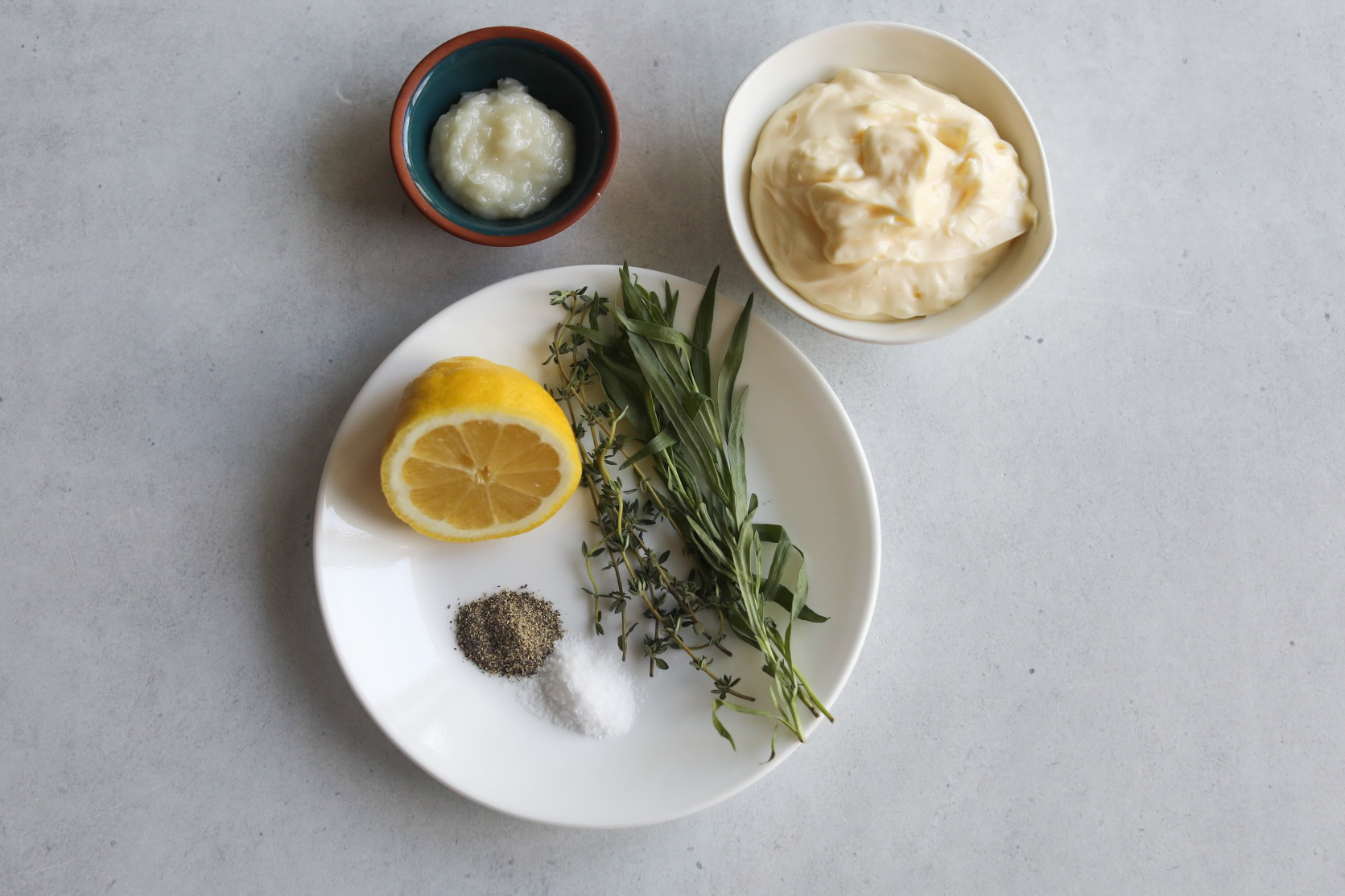 Herb-garlic aiolo sauce ingredients. On a small white plate; salt and pepper, half of a lemon, fresh tarragon and thyme. In a small white bowl is mayonnaise. A small bowl is also filled with garlic paste.