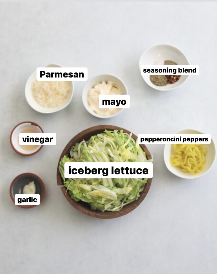 Flat lay of recipe ingredients for Italian Grinder. In small white bowls, seasoning blend, parmesan, mayo, pepperoncini peppers. In a medium sized wooden bowl is iceberg lettuce. In two small white and grey bowls is garlic and vineagar.
