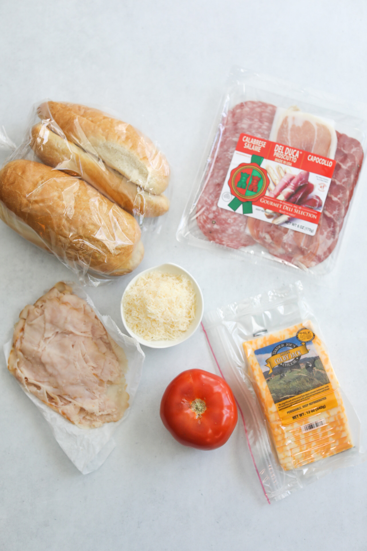Italian Grinder ingredients on a flat lay. Italian meets packaged, crusty bread in a clear bag, white bowl filled with grated parmesan cheese, shaved turkey on parchment paper, a whole tomato and packaged sliced colby jack cheese.